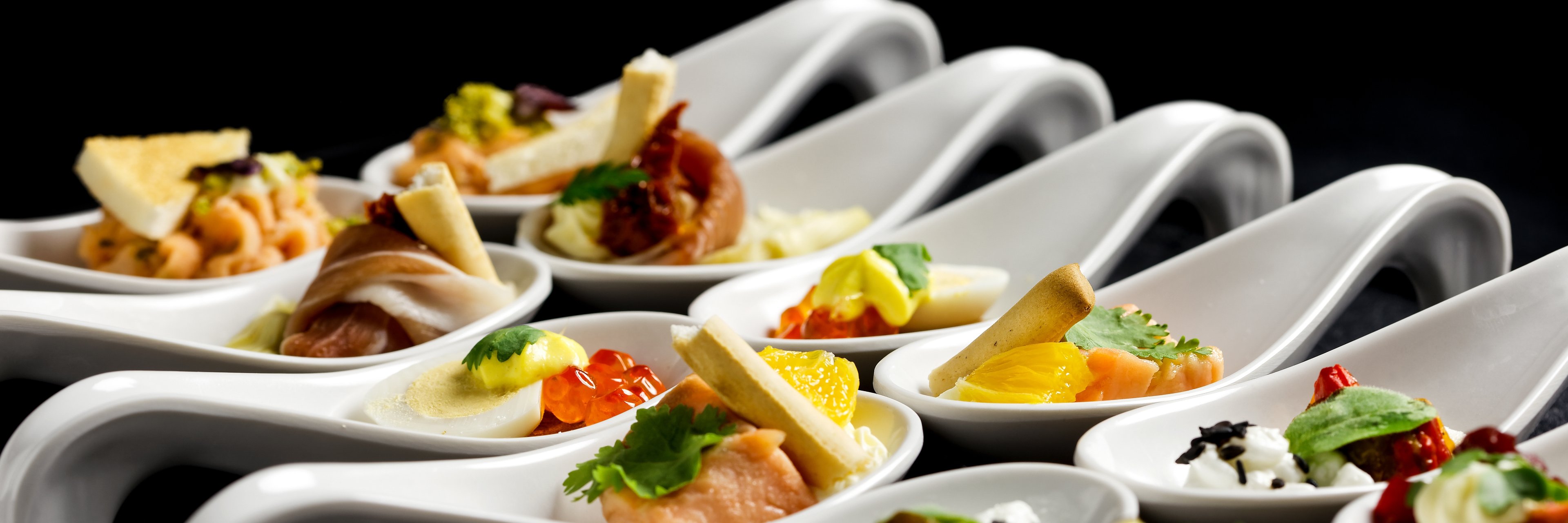 Two rows of canapes on white ceramic spoons. Black background.